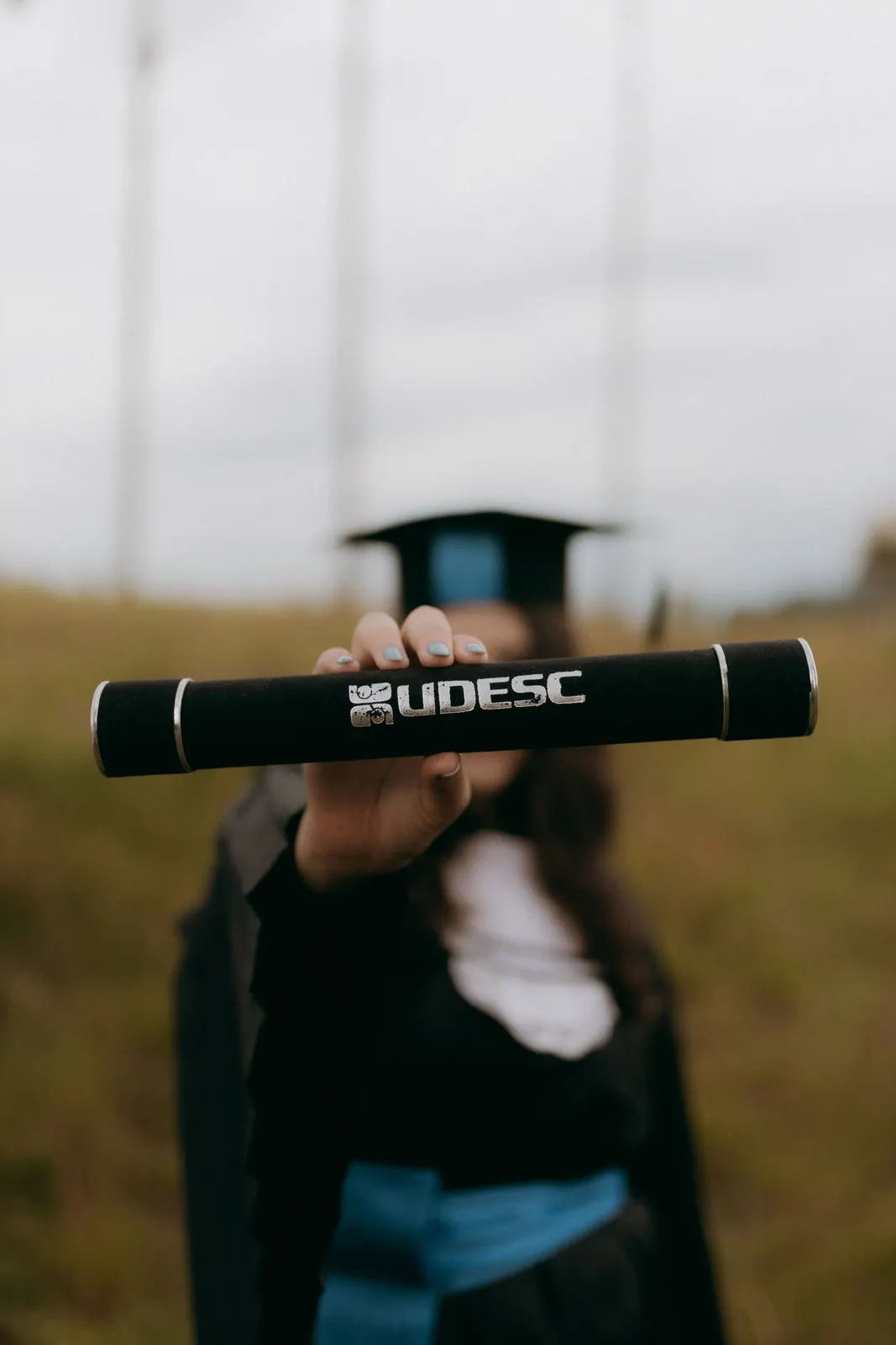 A graduate in cap and gown holding a diploma tube toward the camera (sample image)